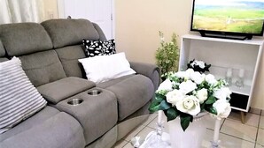 Un salon avec un canapé gris, un vase blanc orné de fleurs, un téléviseur posé sur un meuble et une plante.