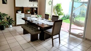 Un coin repas avec une grande table dressée pour quatre personnes, entourée de chaises. À gauche, on trouve un coin cuisine avec des placards et un comptoir. La pièce est dotée d’une grande porte-fenêtre coulissante qui donne sur un patio où l’on aperçoit un hamac et de la verdure.