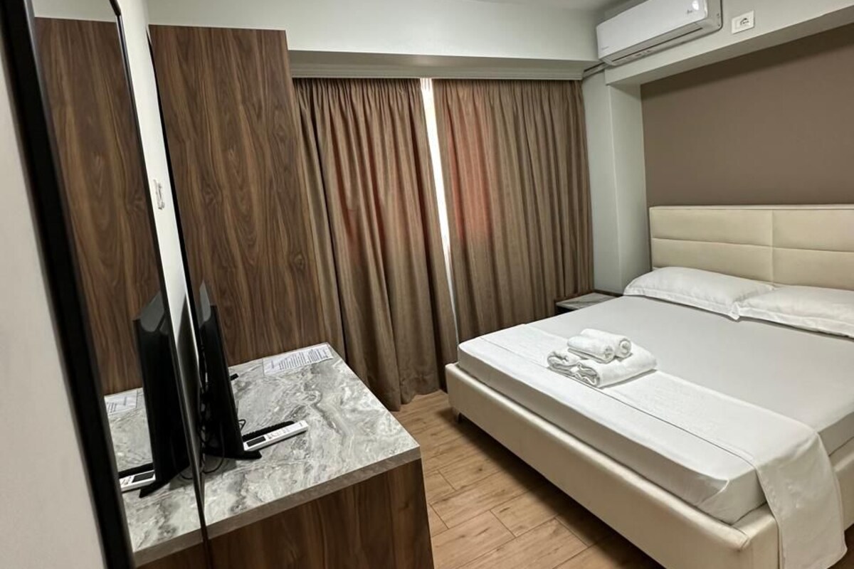 A hotel room with a bed, a bedside table with a flat-screen TV, a mirror, and an air conditioning unit.