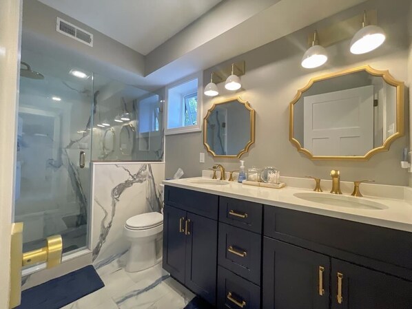 A modern bathroom with a glass shower enclosure, marble backsplash, dual sinks with gold fixtures, and a large mirror with a gold frame.