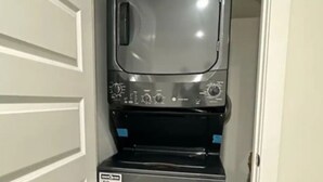 Un appareil compact combinant lave-linge et sèche-linge, en acier inoxydable, installé dans un couloir étroit, avec une porte blanche sur le côté gauche.