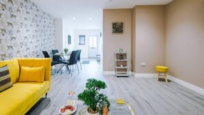 Een moderne woonkamer met een gele bank, een eethoek met stoelen, een kleine tafel en een plant.