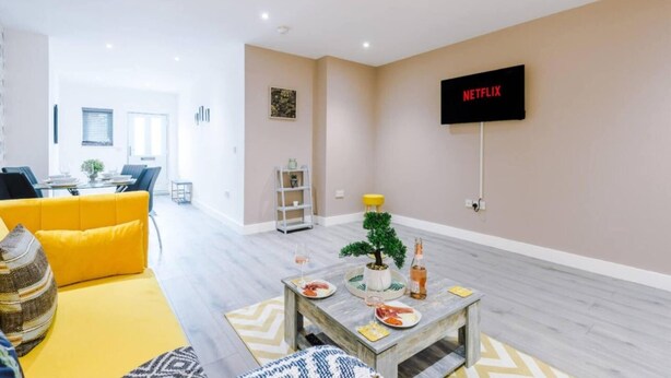 Een moderne woonkamer met een gele bank, een bijzettafeltje met een plant en drankjes, een eethoek op de achtergrond en een aan de muur bevestigde televisie met de tekst 'NETFLIX'.