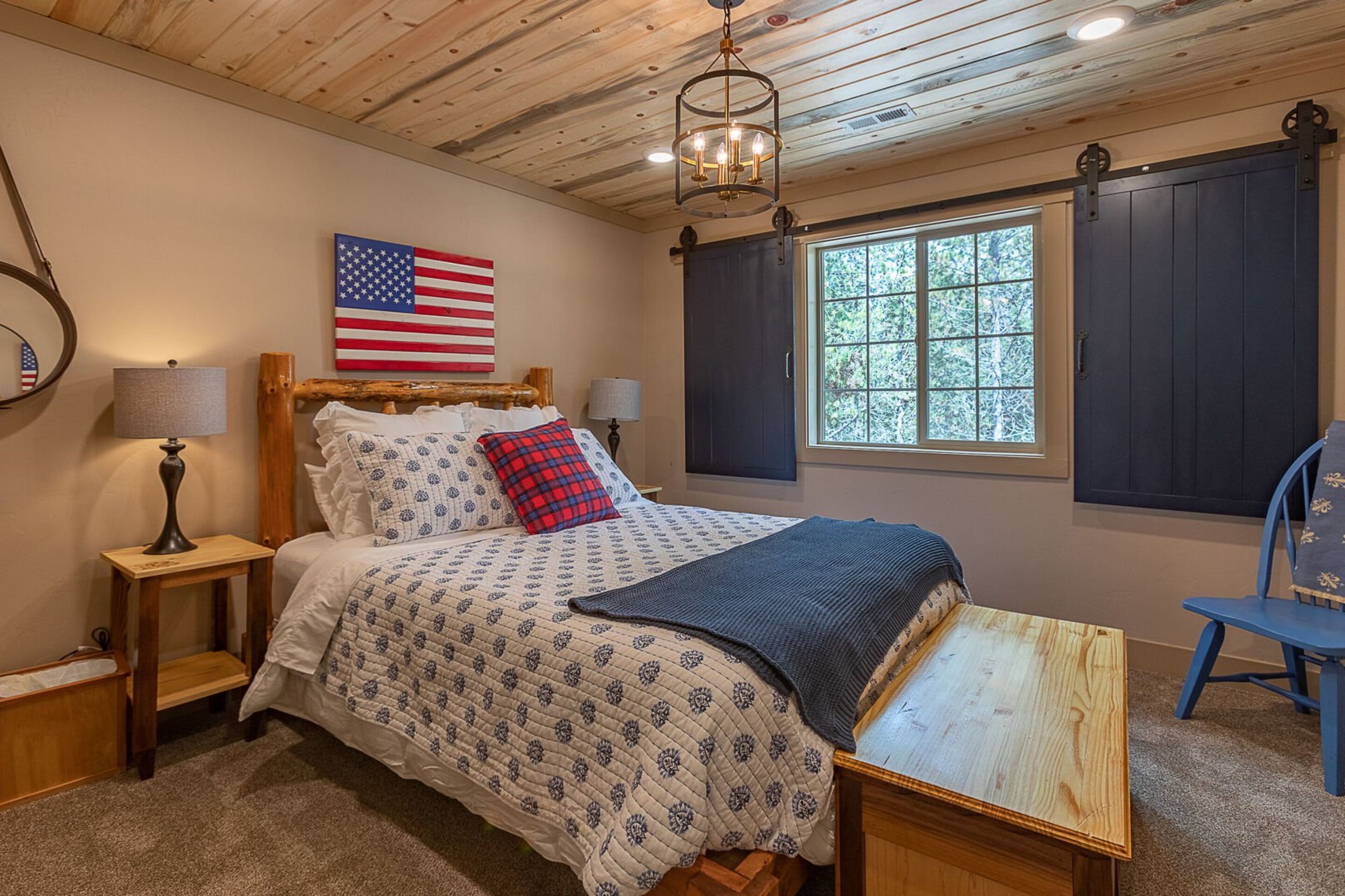 Un dormitorio con techo de madera, una cama con ropa de cama estampada, una mesita, una silla, una lámpara, una ventana con contraventanas y una bandera estadounidense en la pared.