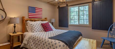 Un dormitorio con techo de madera, una cama con ropa de cama estampada, una mesita, una silla, una lámpara, una ventana con contraventanas y una bandera estadounidense en la pared.