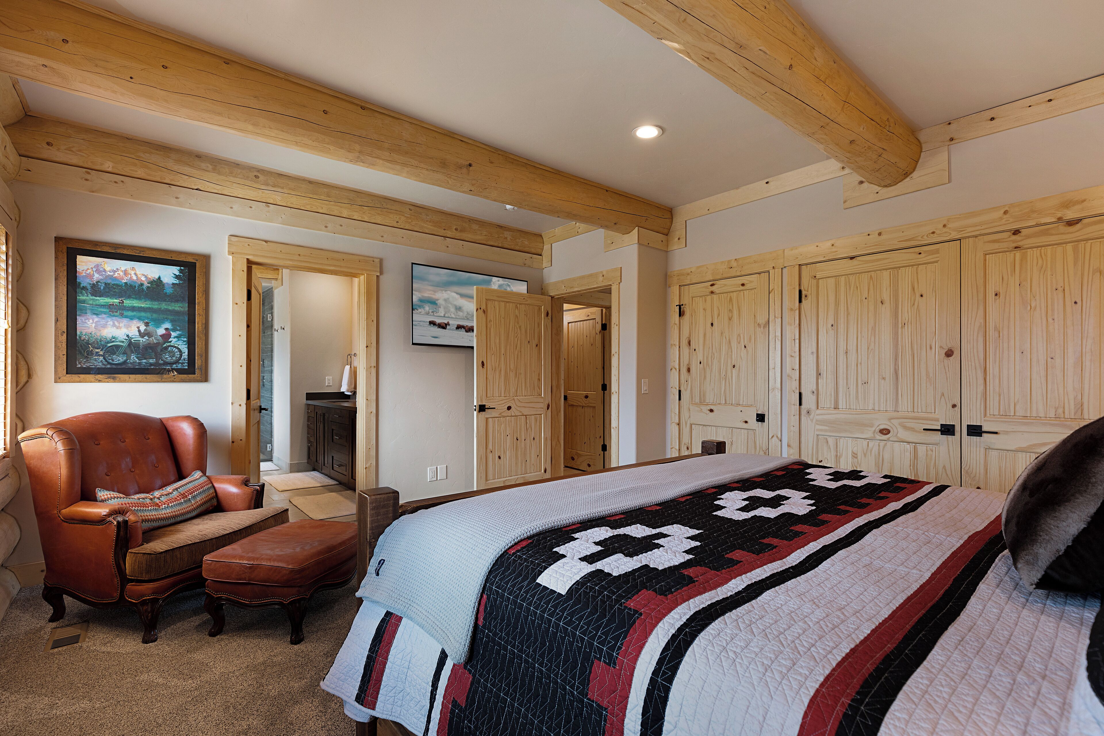 A bedroom with a large bed, wooden ceiling beams, a leather armchair, and a view of the countryside through the window.