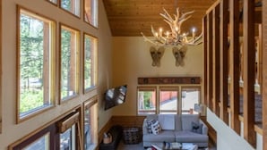L’intérieur chaleureux d’un chalet avec un plafond en bois, de grandes fenêtres, une cheminée et un téléviseur fixé au mur.