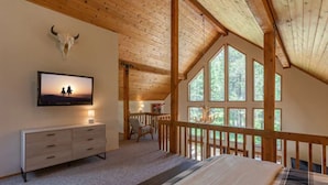 Une chambre avec un plafond en bois, un téléviseur fixé au mur, un lit recouvert d’une couverture à carreaux et une grande fenêtre donnant sur la verdure.