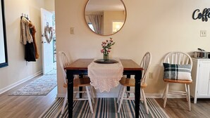 A dining area with a wooden table, two chairs, a lace table runner, a mirror, and a wall-mounted sign.