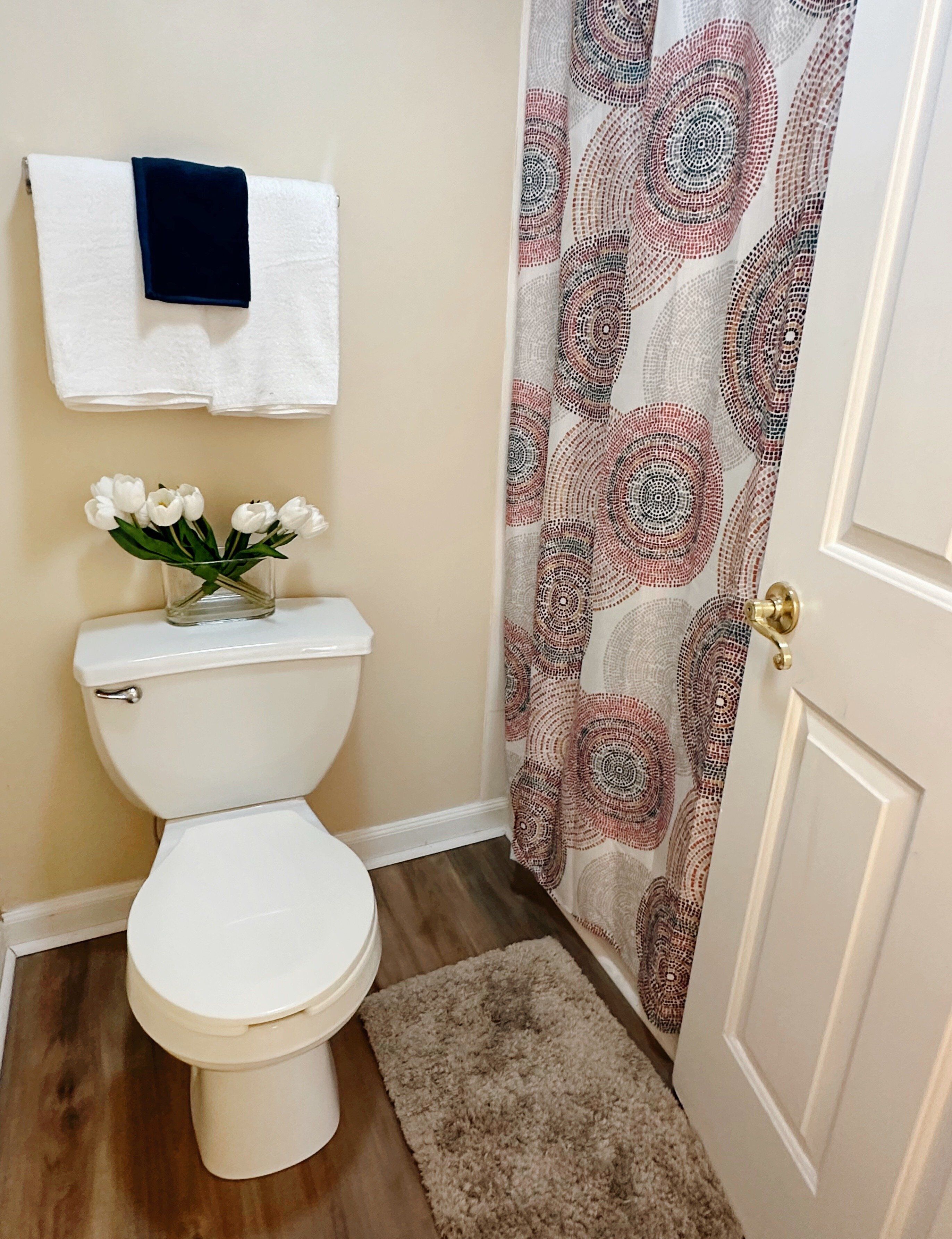 Een badkamer met een toilet, een witte handdoek aan de muur, een vaas met witte bloemen en een badgordijn met een patroon.