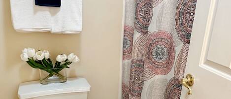 A bathroom with a toilet, a white towel hanging on the wall, a vase with white flowers, and a patterned shower curtain.