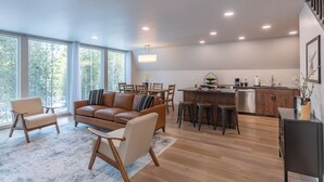 Ein modernes Wohnzimmer mit einer braunen Ledersofa, einem Holztisch und einem Essbereich mit Holzstühlen. Im Hintergrund ist eine Küche mit dunkler Arbeitsplatte und Edelstahlgeräten zu sehen.