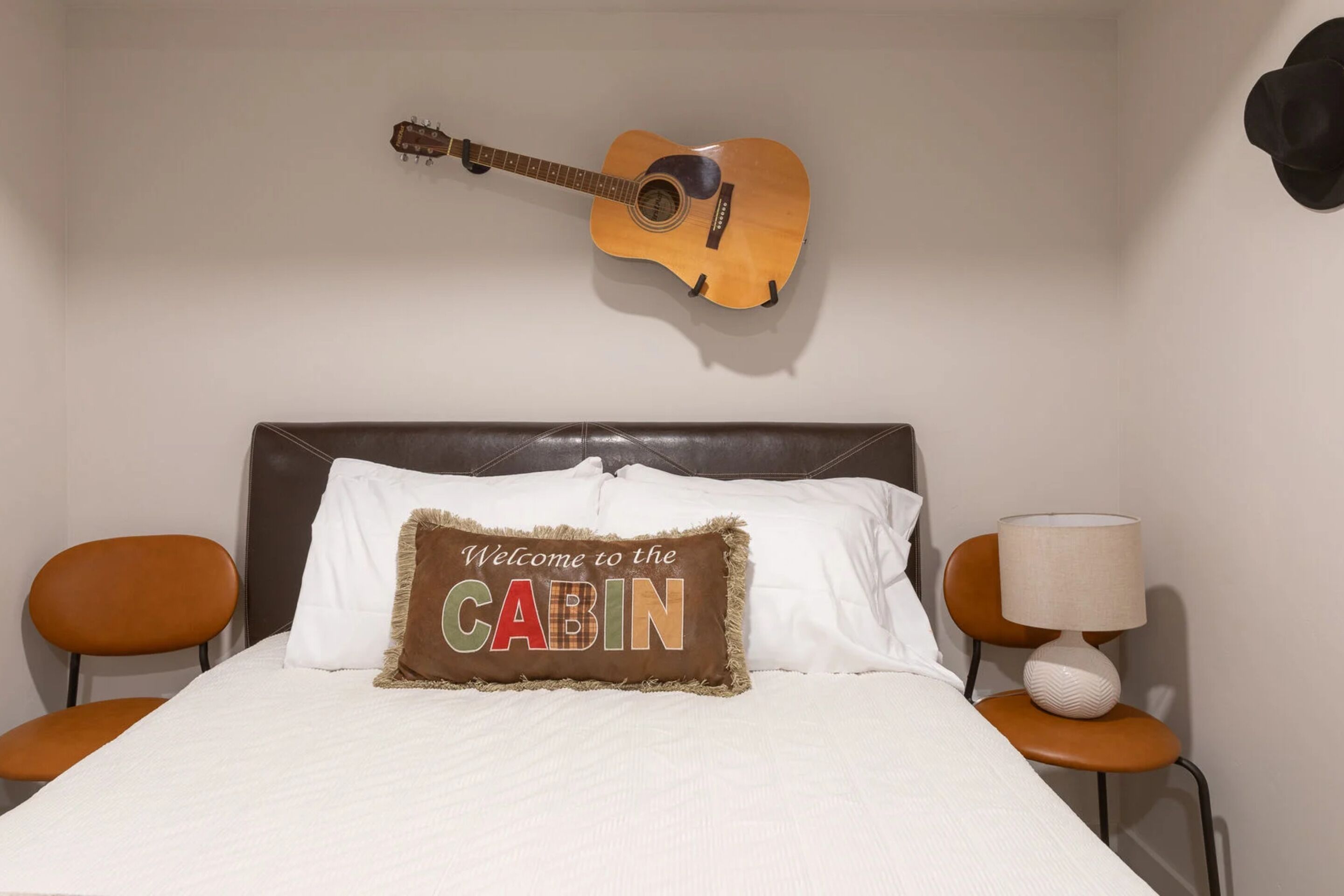 Un lit avec un oreiller sur lequel est brodé le mot « BIENVENUE AU CABIN », une guitare accrochée au mur et deux chaises, dont l’une est surmontée d’une lampe.