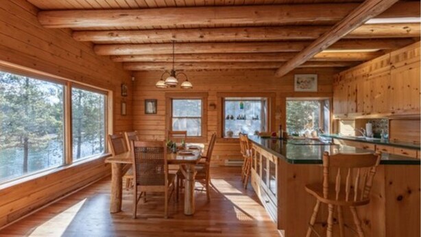 A wooden cabin interior with a dining area, kitchen, and large windows offering views of the forest. - Buffalo Bliss (Island Park)