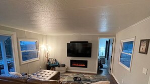 A living room with a large leather sofa, a flat-screen TV mounted on the wall, a fireplace, and a window with a view of the outdoors.