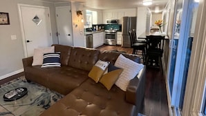 A living room with a brown sectional sofa, a dining area with a table and chairs, a kitchen with appliances, and a hallway leading to a door.