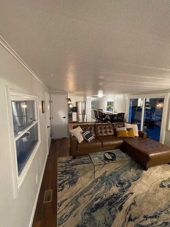 A compact living space with a brown leather sofa, a glass coffee table, and a patterned rug. A kitchen area is visible in the background with modern appliances.