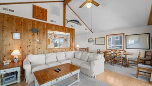 A cozy living room with a sectional sofa, wooden ceiling beams, a ceiling fan, and a dining area with wooden chairs and a table.