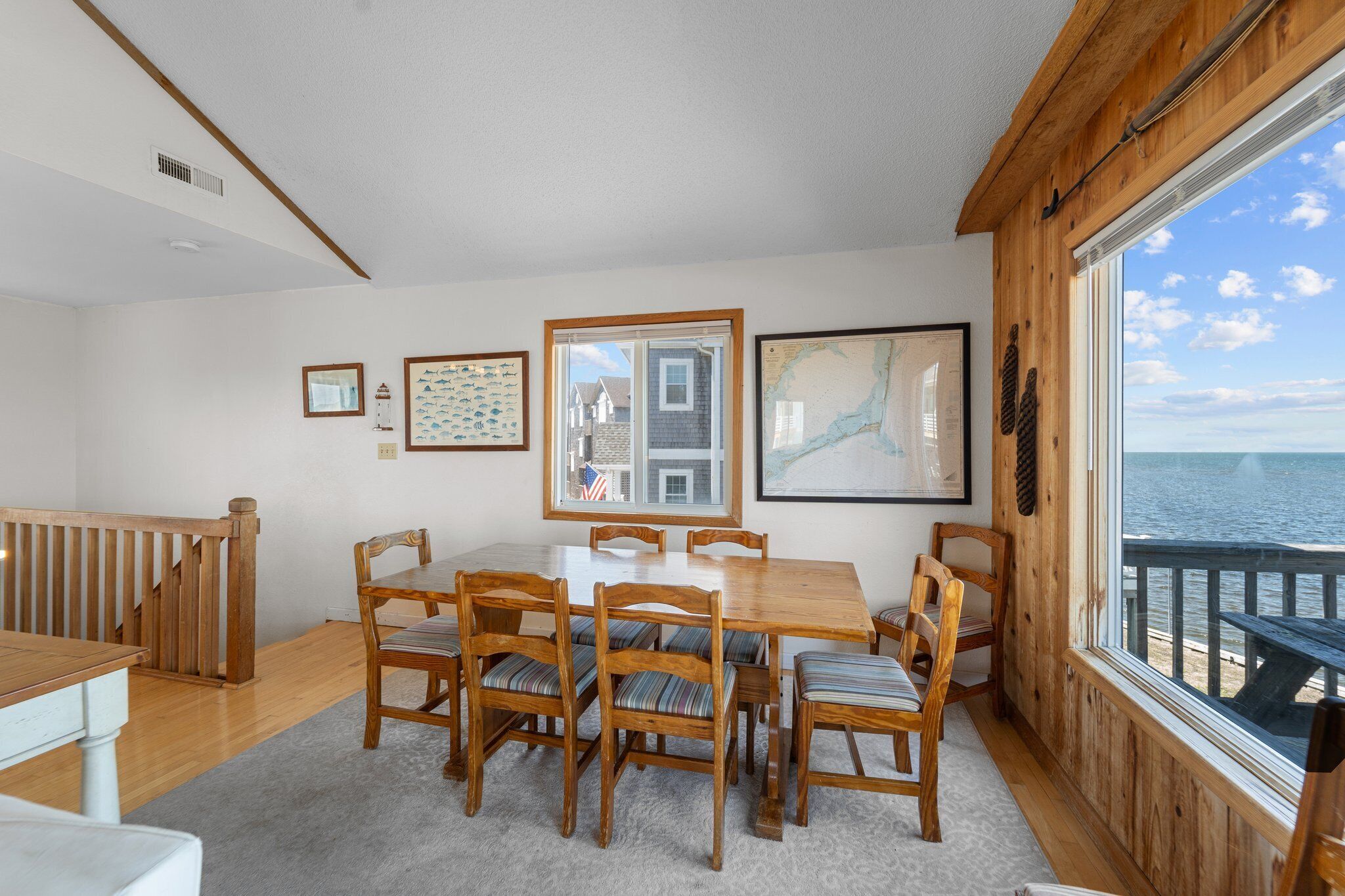 A dining area with a wooden table and chairs, framed pictures on the wall, and a view of the ocean through a large window.