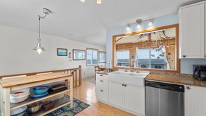 A modern kitchen with a large window, wooden cabinets, a dishwasher, and a dining area with a wooden table and chairs.