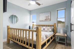 Ein Schlafzimmer mit einem hölzernen Bett, einem runden Spiegel, einem Nachttisch und einem Fenster mit Blick auf eine Strandlandschaft.