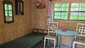 Una habitación de madera con una cama de metal, una mesita y sillas. Hay estanterías con libros y una ventana con vistas a zonas verdes.