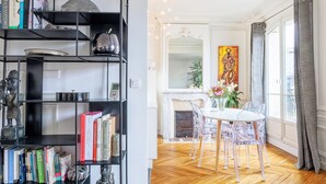 Una stanza con pavimento in parquet a spina di pesce, un camino bianco e un tavolo da pranzo con sedie trasparenti. Si nota una libreria con libri e oggetti decorativi.