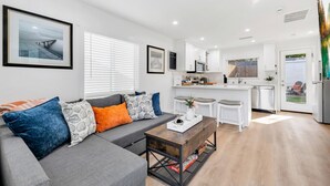 Een moderne woonkamer met een grijze bank, een salontafel en een keuken met witte kasten en een magnetron.