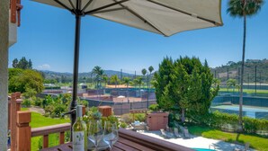 A wooden table with a bottle of wine and glasses, an umbrella, and a view of a tennis court and greenery.