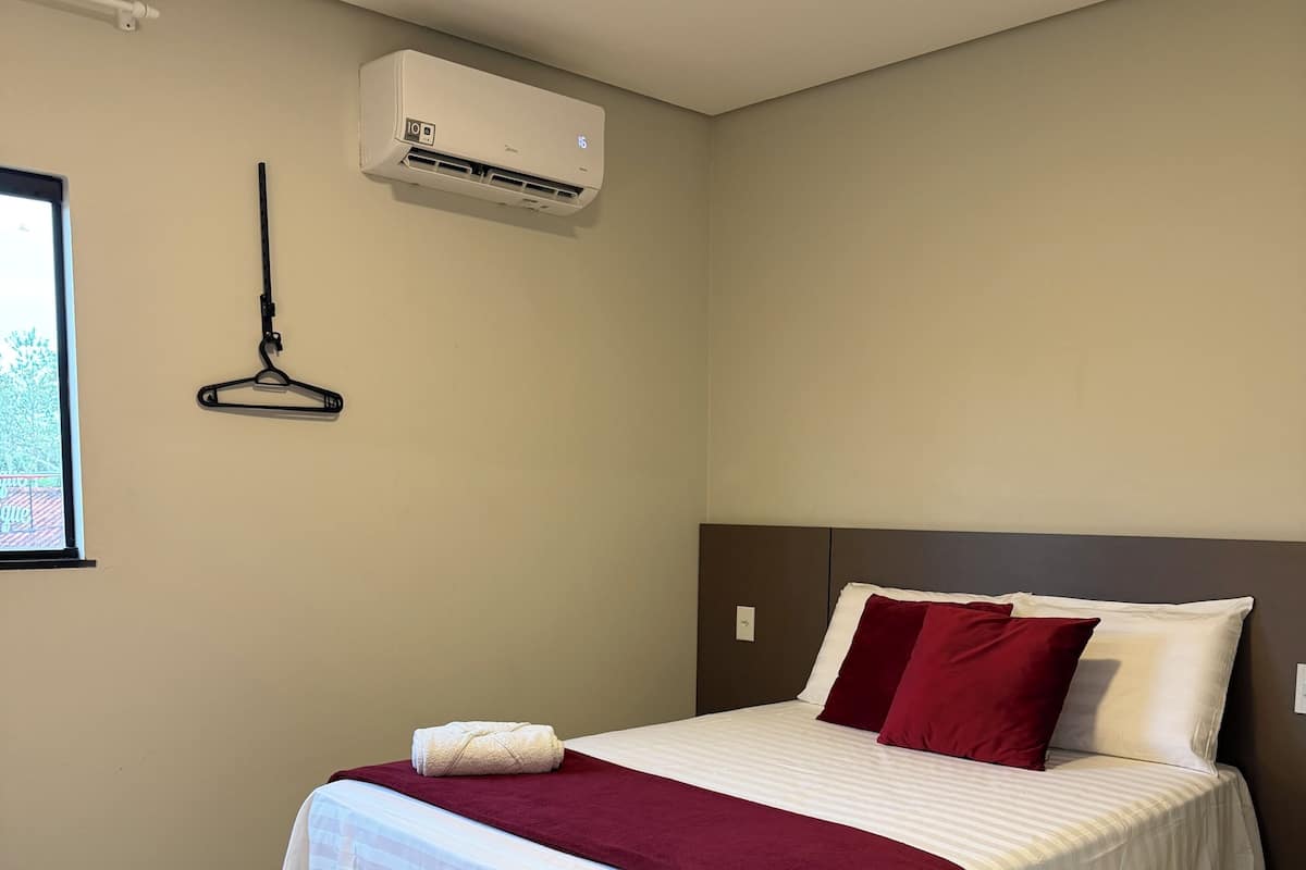 Una cama bien hecha con una colcha blanca y roja, un perchero negro colgante y un equipo de aire acondicionado en la pared.