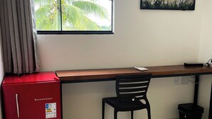 Een kamer met een bureau, een stoel, een rode koelkast, een raam met uitzicht op palmbomen en een aan de muur hangend schilderij.