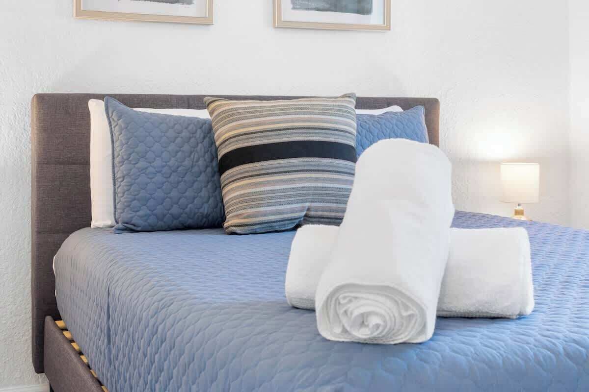 Una cama bien hecha con colcha azul, dos almohadas y toallas blancas enrolladas.