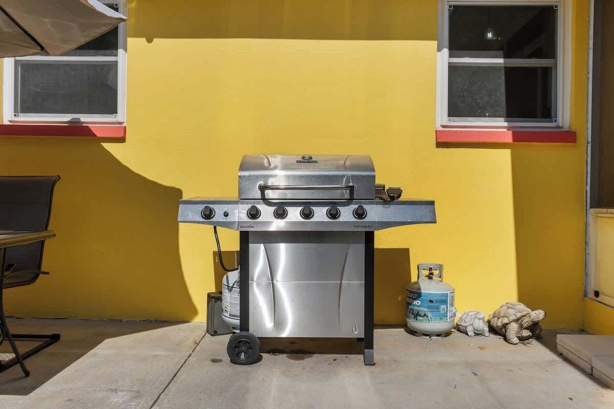 Un barbecue au gaz portatif, muni de roulettes, avec une bouteille de propane et une petite statue de tortue, placé sur une terrasse en béton devant un mur jaune orné de fenêtres.
