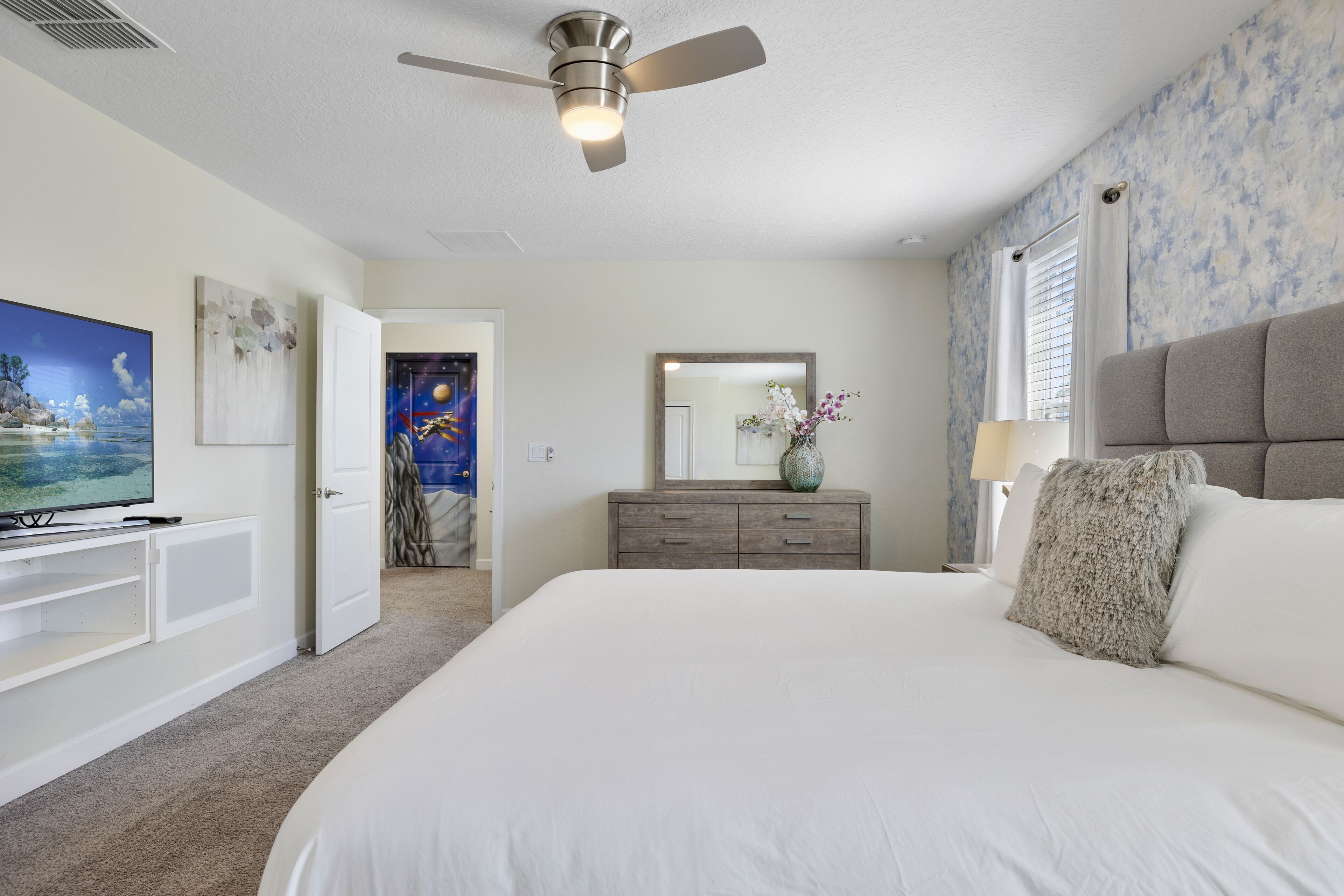 A bedroom with a large bed, a ceiling fan, a dresser, a TV, and a door leading to another room.
