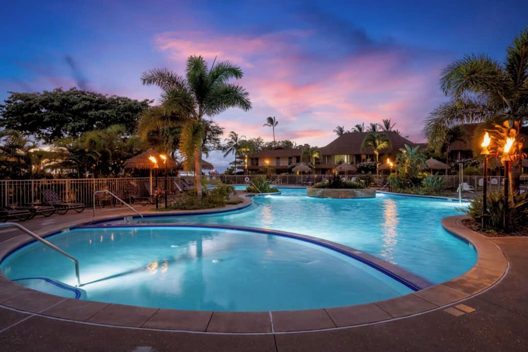 Ein Pool mit geschwungener Form, umgeben von Palmen und Liegestühlen, mit Gebäuden im Hintergrund.
