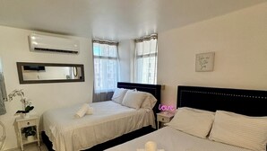 A hotel room with two beds, a mirror, an air conditioning unit, a bedside table with a plant, and a wall-mounted sign.
