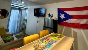 A living room with a dining area, a flat-screen TV mounted on the wall, a flag, and a table with a colorful tablecloth.