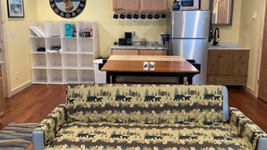 A compact kitchen with wooden cabinets, a stainless steel refrigerator, and a coffee maker. There is a dining table with two cups and a patterned sofa in the foreground. - Wapiti Cabin - a Wolf Crossing Cabin next to Yellowstone (Silver Gate)
