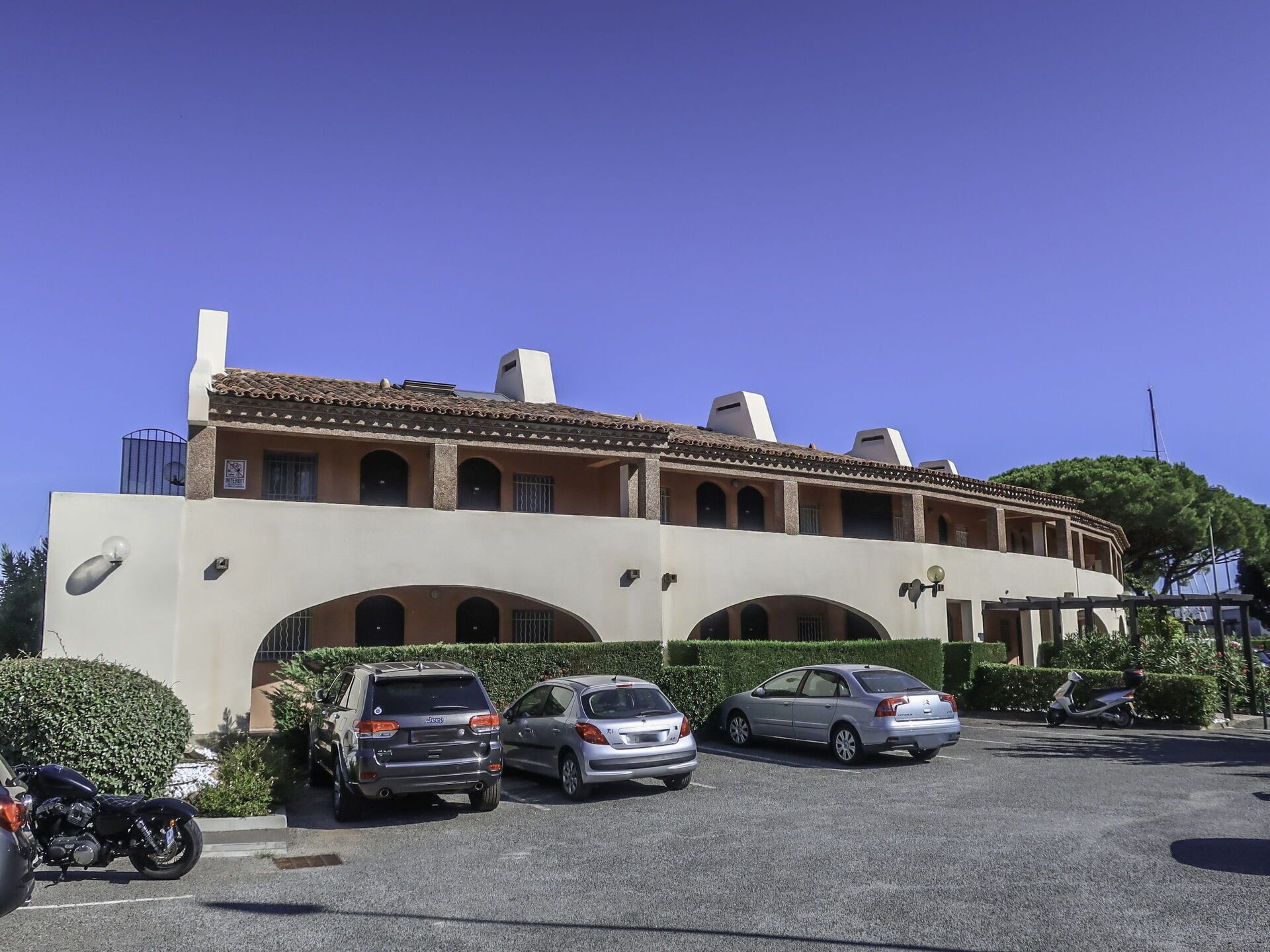 A building with a beige facade, multiple arches, and a tiled roof. There are three cars parked in front and a motorcycle to the left.