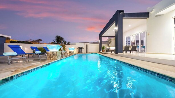 A swimming pool with lounge chairs, a modern building with glass doors, and a sunset sky. - Modern & Spacious 4BR3BA w/ Pool & Close to Beach! (Noord)