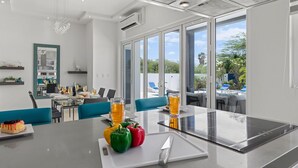 A modern kitchen with a glass dining table, a built-in stove, and a view of the outdoor pool area. - Modern & Spacious 4BR3BA w/ Pool & Close to Beach! (Noord)
