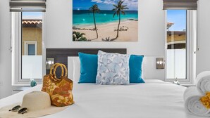 A hotel room with a bed, two pillows, a beach-themed painting, a window with a view of the beach, and a sun hat on the bed. - Modern & Spacious 4BR3BA w/ Pool & Close to Beach! (Noord)