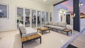 A modern living room with a sofa, two coffee tables, and a sliding glass door leading to a balcony. - Modern & Spacious 4BR3BA w/ Pool & Close to Beach! (Noord)