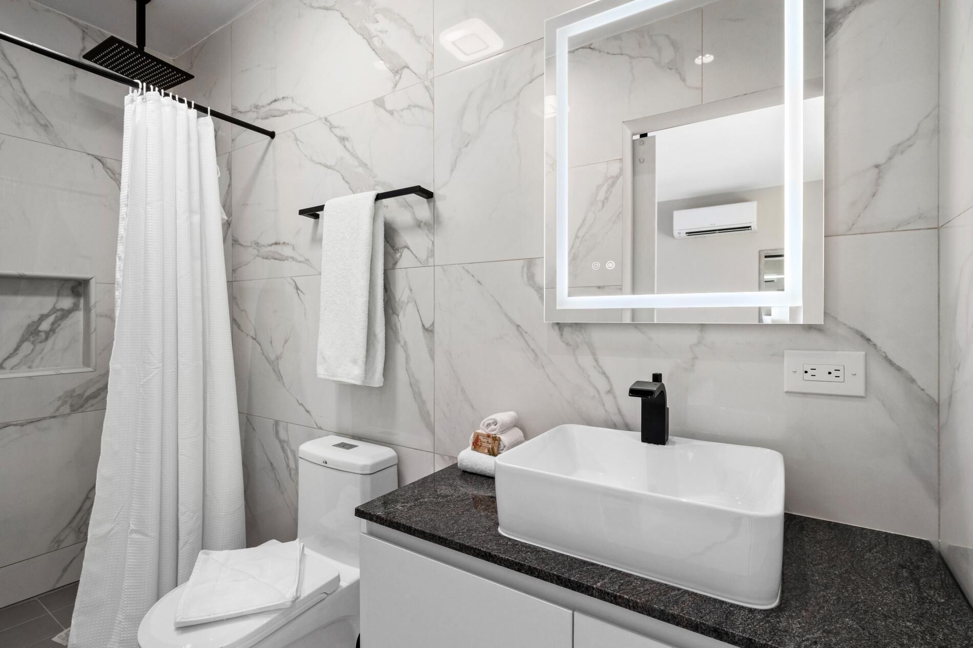 Ein modernes Badezimmer mit Marmorwand, Duschvorhang, Toilette, Waschbecken inklusive Spiegel und Handtuchhalter.