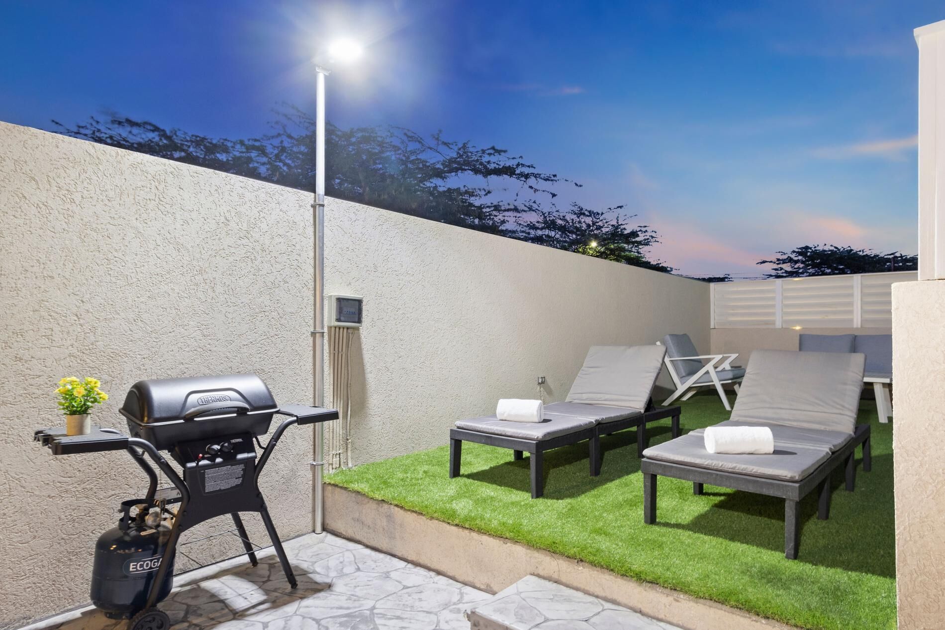 Een terras buiten met een barbecue, lounge fauteuils en een kleine tafel.