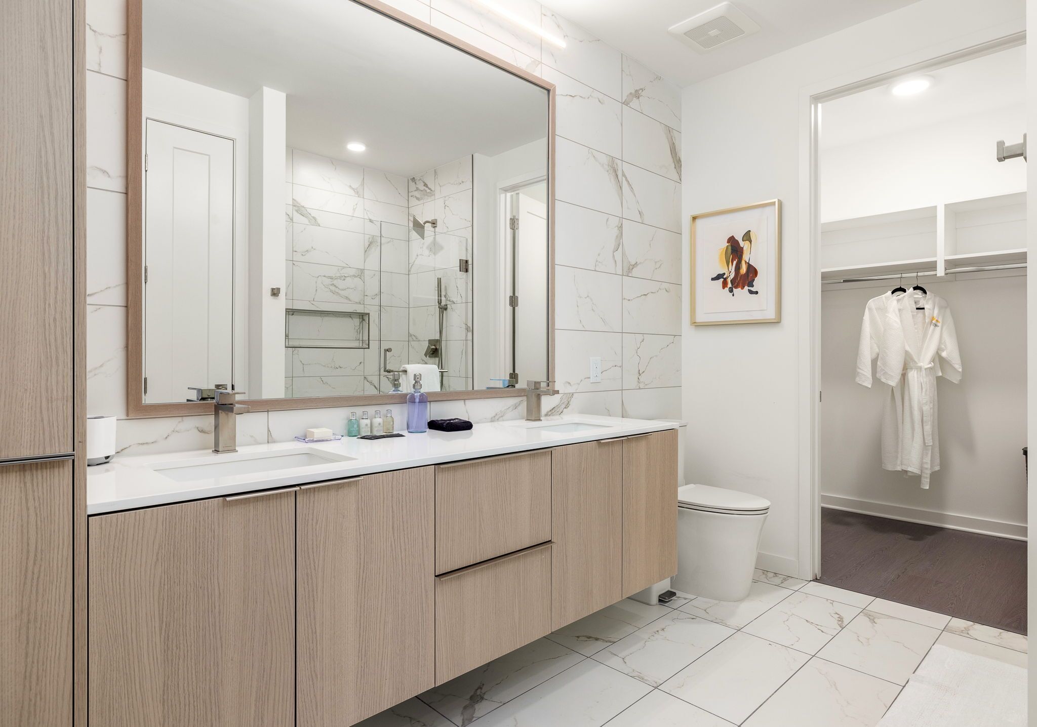 A modern bathroom with a large mirror, two sinks, a shower, and a toilet.