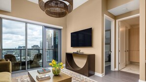 A modern living room with a large window, a flat-screen TV, a curved coffee table, and a view of the city. - 26th Floor - Living Daylight (Atlanta)