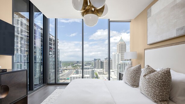 A modern hotel room with a large bed, a desk, and a view of the cityscape through floor-to-ceiling windows. - 26th Floor - Living Daylight (Atlanta)