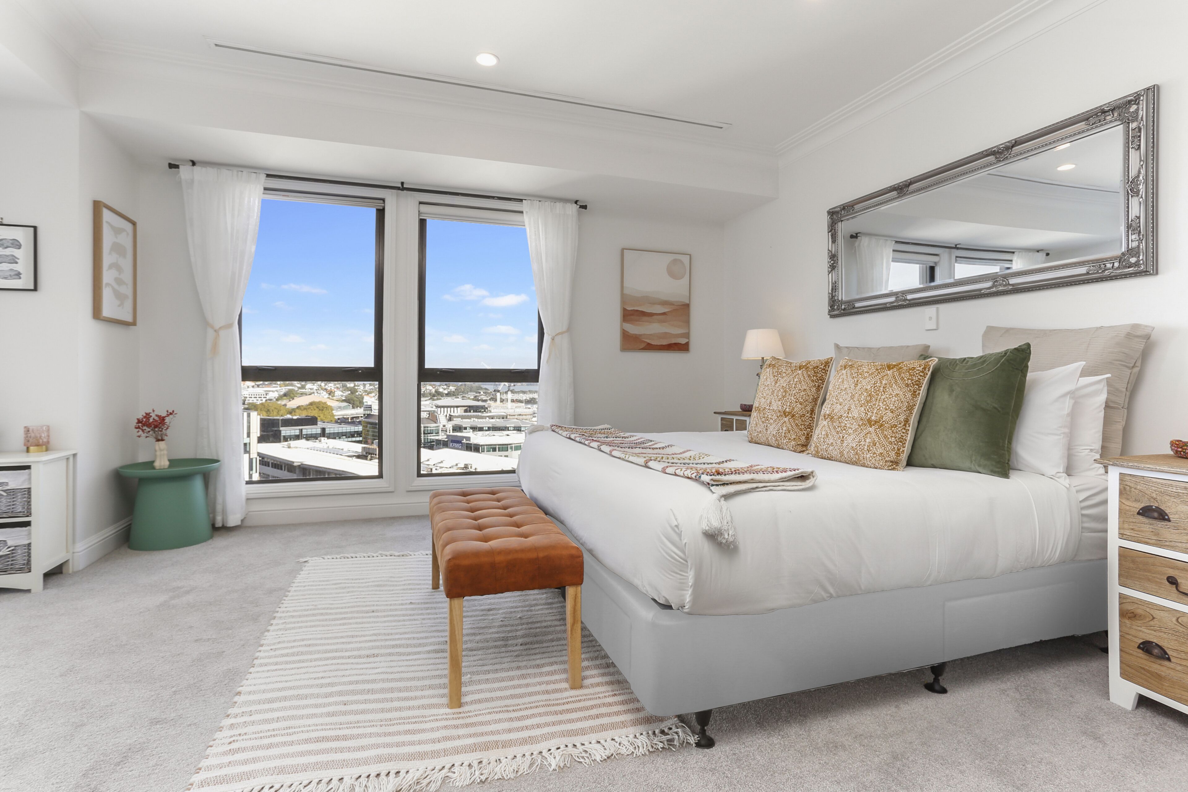 A bedroom with a large bed, a nightstand, a dresser, a window with curtains, a view of buildings, and a mirror on the wall.