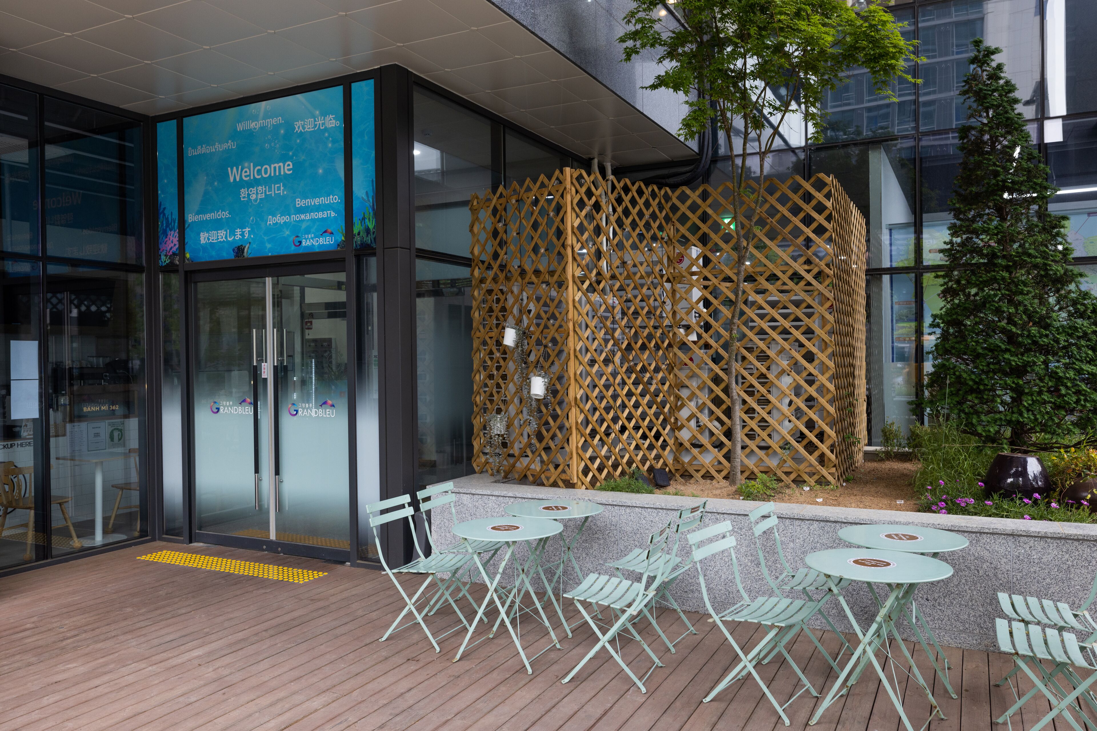 A modern building entrance with a welcome sign, glass doors, and outdoor seating with chairs and tables.